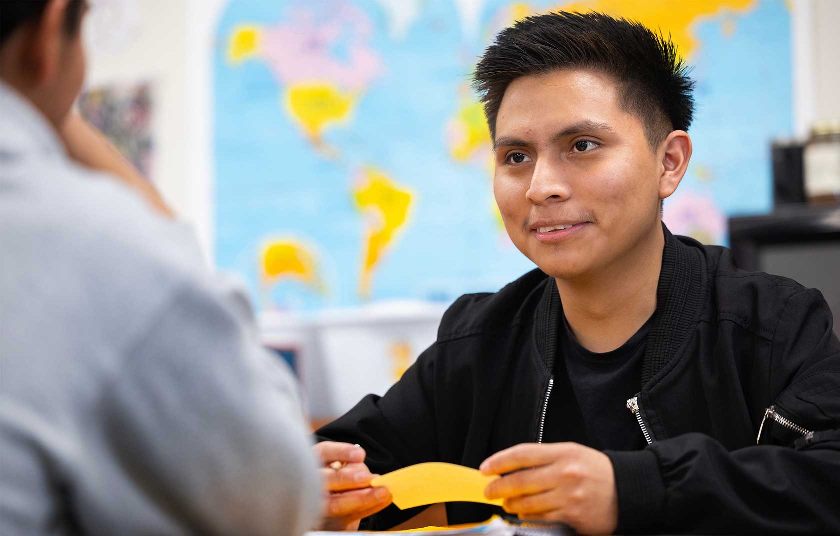 A young male student in a black jacket holds orange paper while engaged in a conversation in a classroom with a world map in the background.