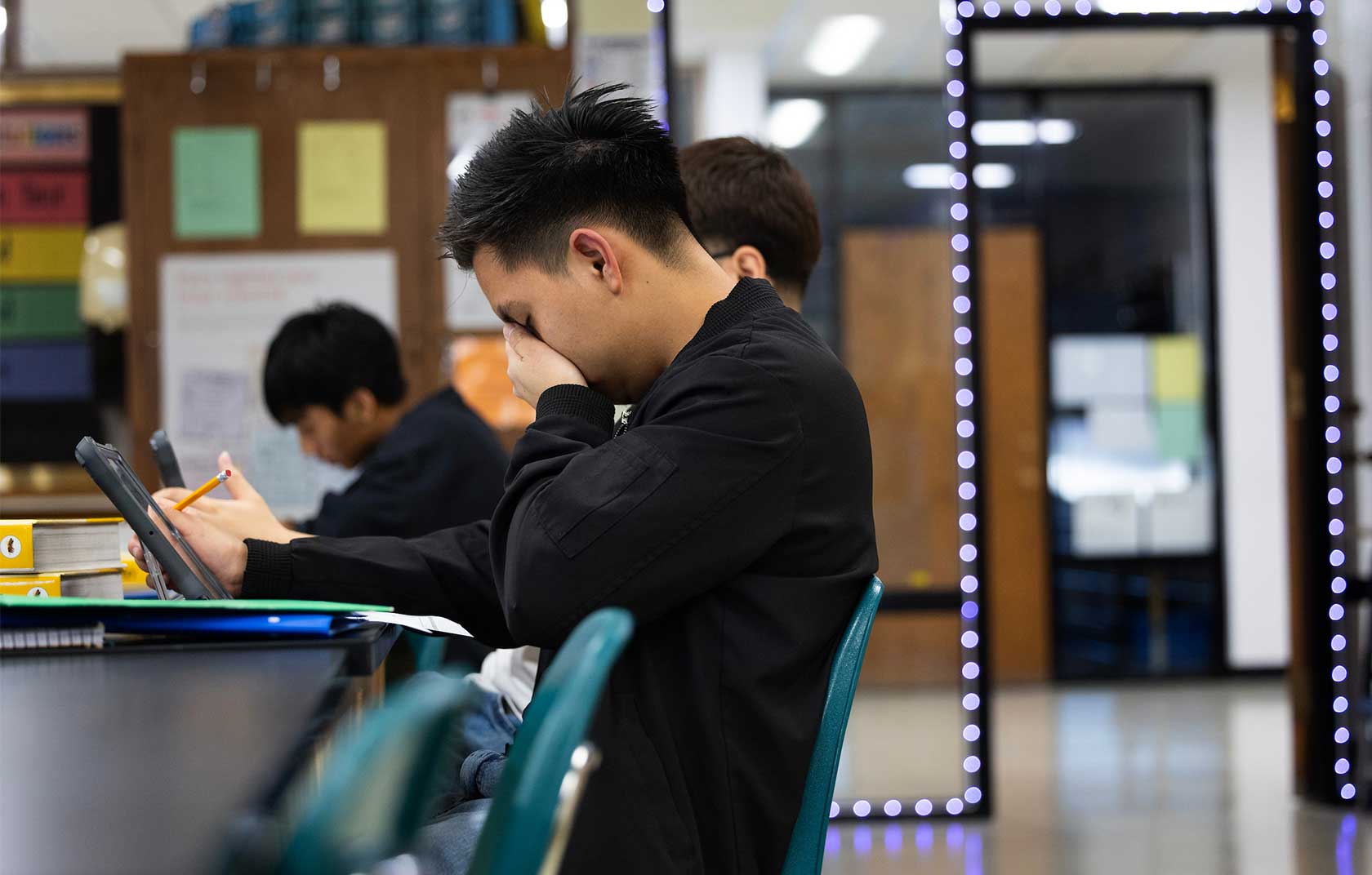 A distressed student in a black jacket holds a tablet and covers his eyes with one hand in a classroom, while other students are engaged with their devices in the background.