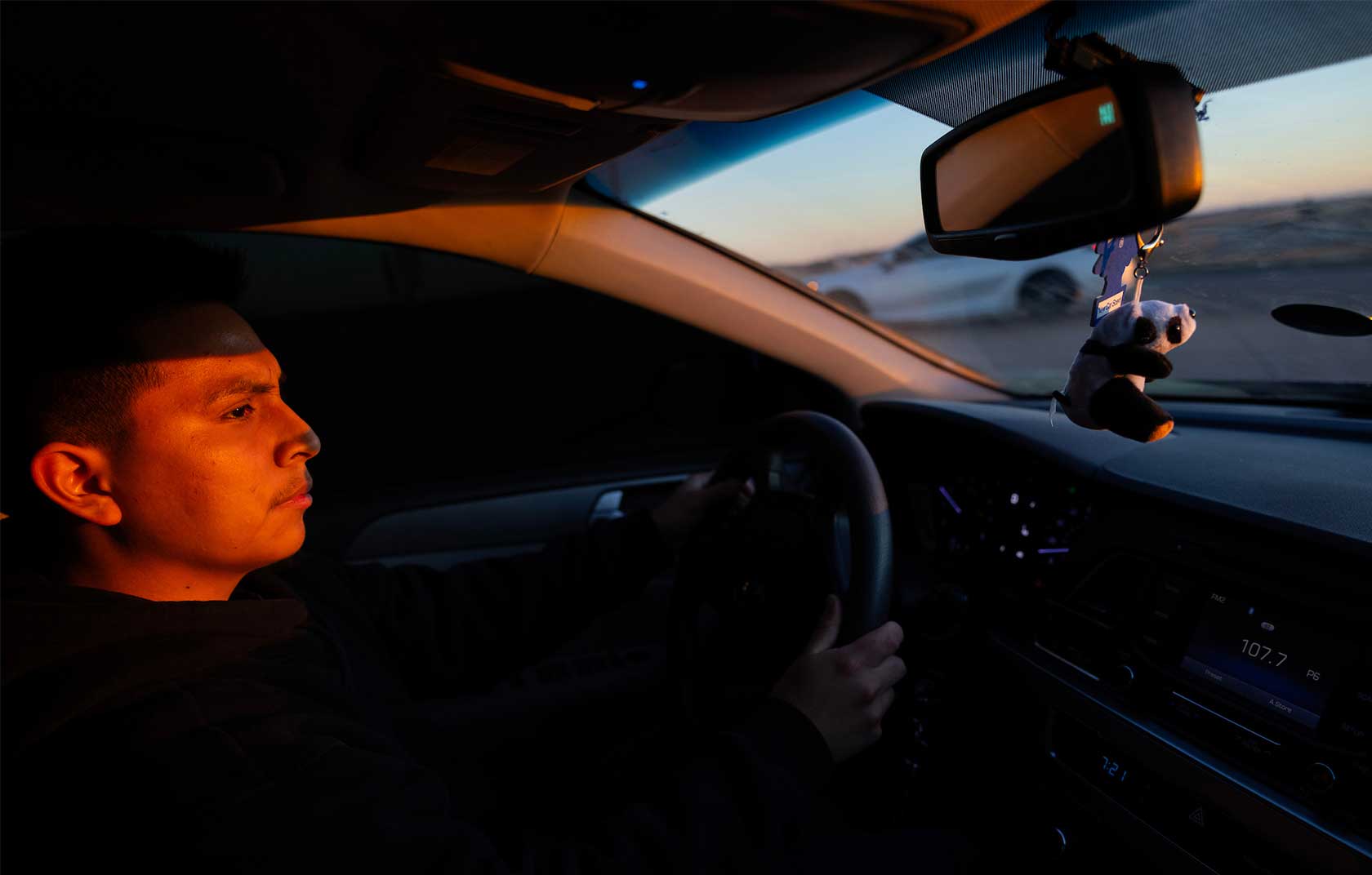 A young man sits in the driver's seat of a car, illuminated by warm sunset light, with a plush panda keychain hanging from the rearview mirror.