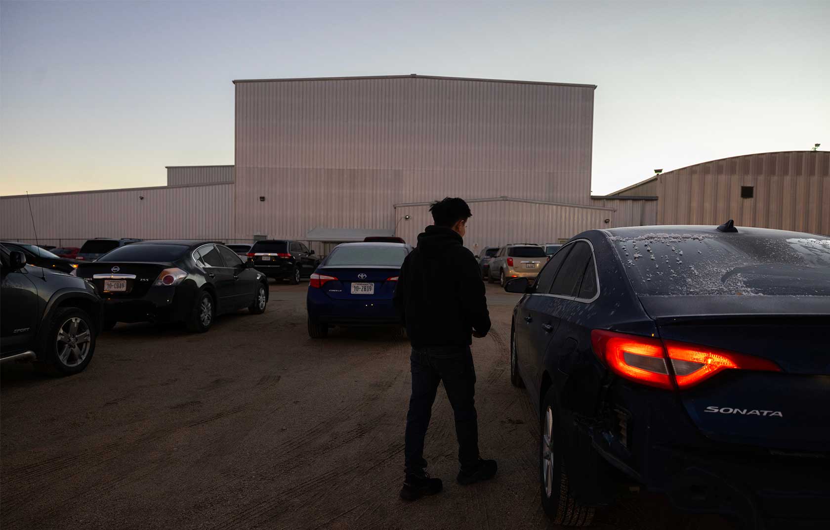 A person stands next to a blue car in a parking lot during twilight, with several other cars and a large industrial building in the background.