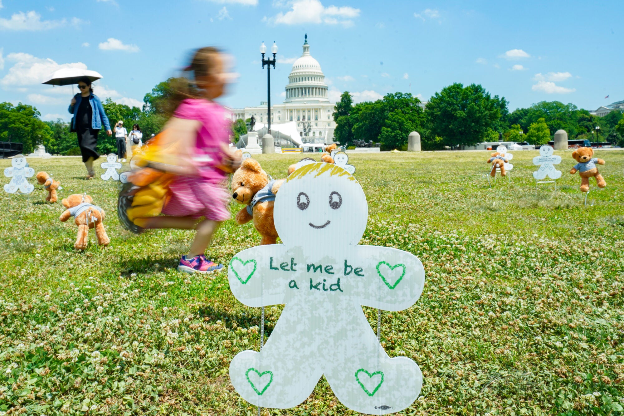 A young girl in a pink dress runs in the grass outside of the U.S. Capitol, where there is a white paper sign shaped like a child with hearts on it that says Let me be a kid. There are other similar signs and teddy bears placed throughout the lawn as part of a vigil. An adult woman is in the background holding an umbrella.