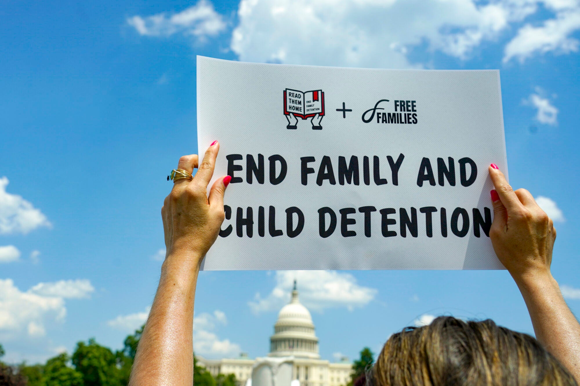 An adult's hands hold up a white sign with a logo for Read Them Home plus Free Families that reads End family and child dentention. The person is holding the sign in front of the U.S. Capitol building.