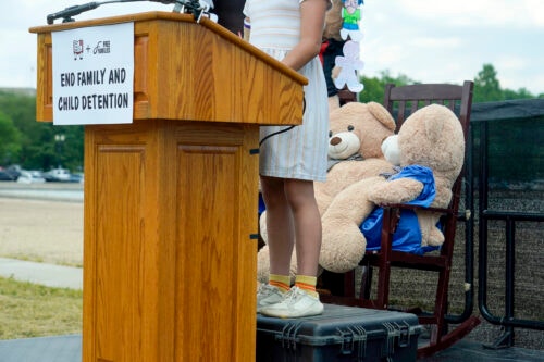 A brown, wooden podium with a sign that says End family and child detention is shown with a child standing behind it on a black bin. The child's head is not pictured as it is out of the frame of the photo. Two large teddy bears are in chairs behind the child.