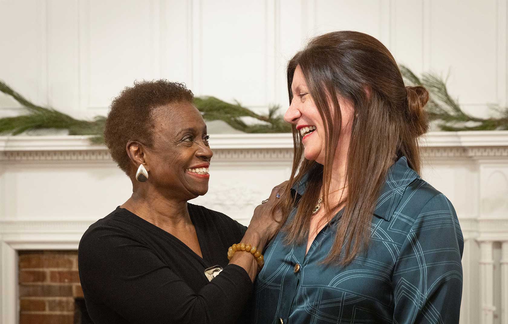 Two women smiling and laughing together in a cozy indoor setting, with greenery on the mantel behind them.