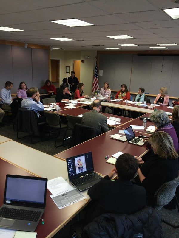 A meeting in a conference room with people seated around a large table, using laptops and taking notes.