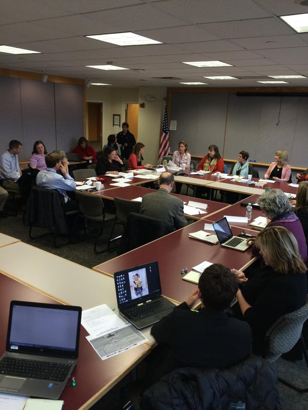 A meeting in a conference room with people seated around a large table, using laptops and taking notes.