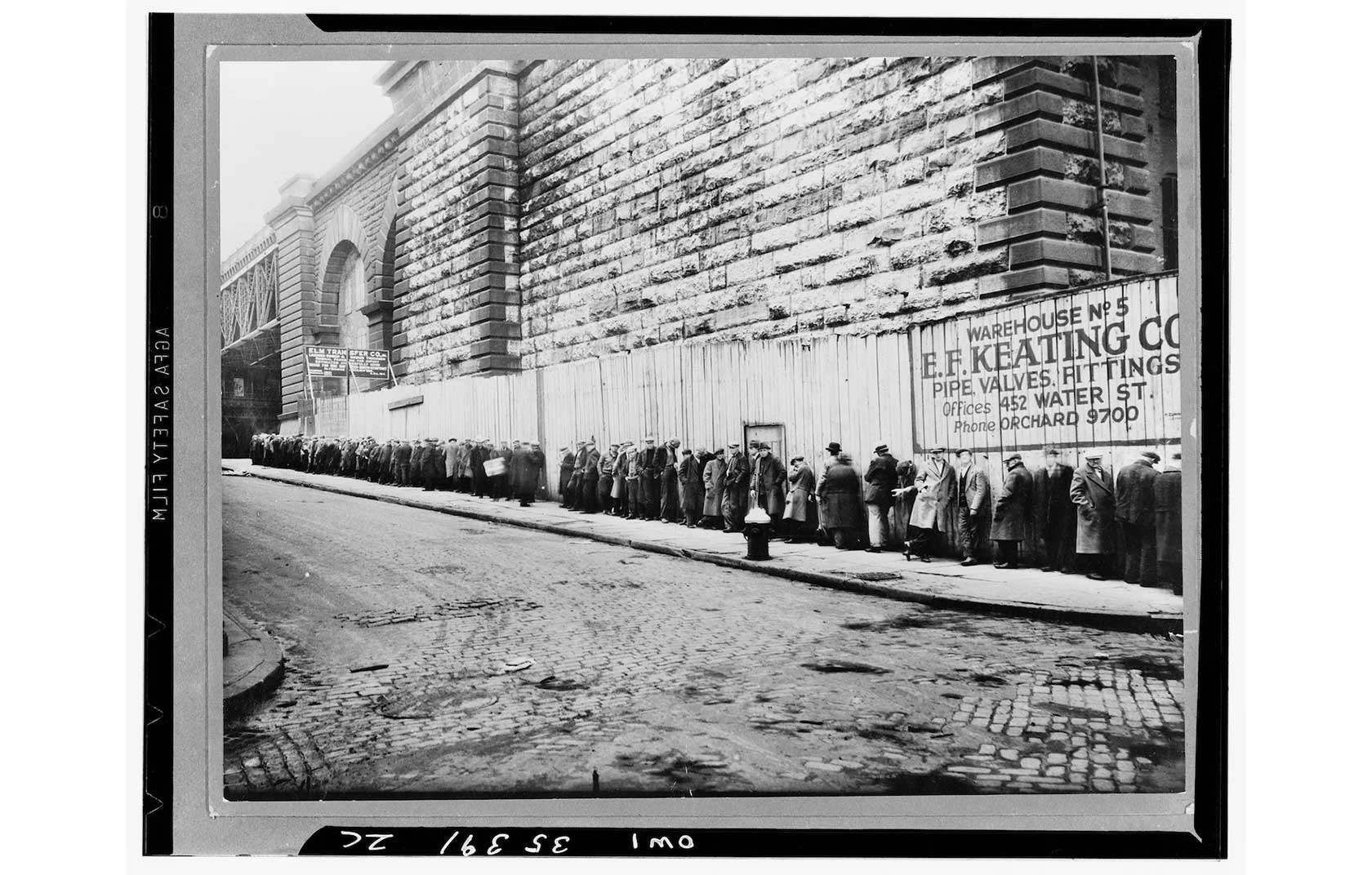 A line of men in early 20th-century clothing waiting along a cobblestone street beside a large stone wall.