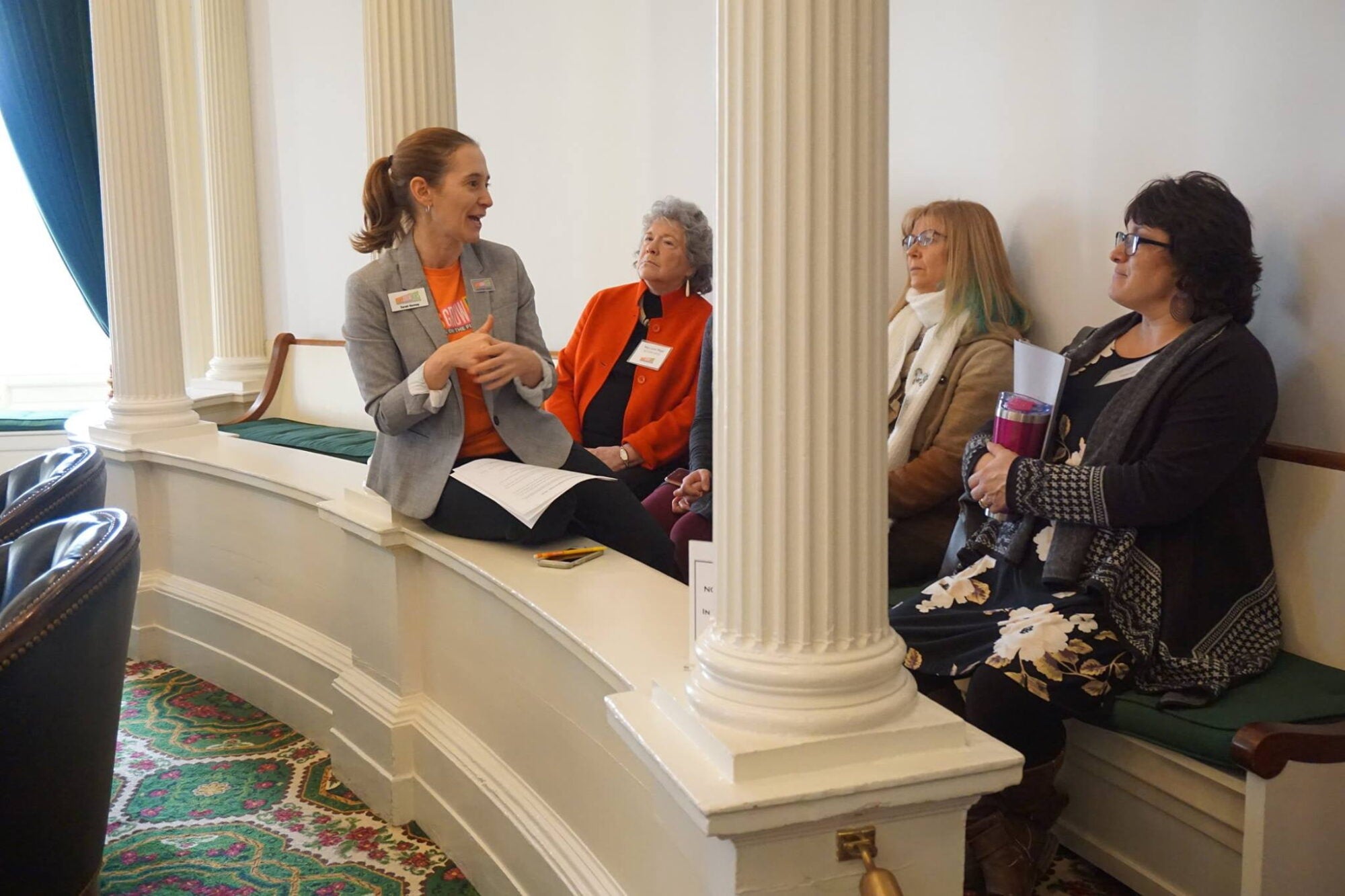 Four women seated in a room with columns, one speaking animatedly, others listening.