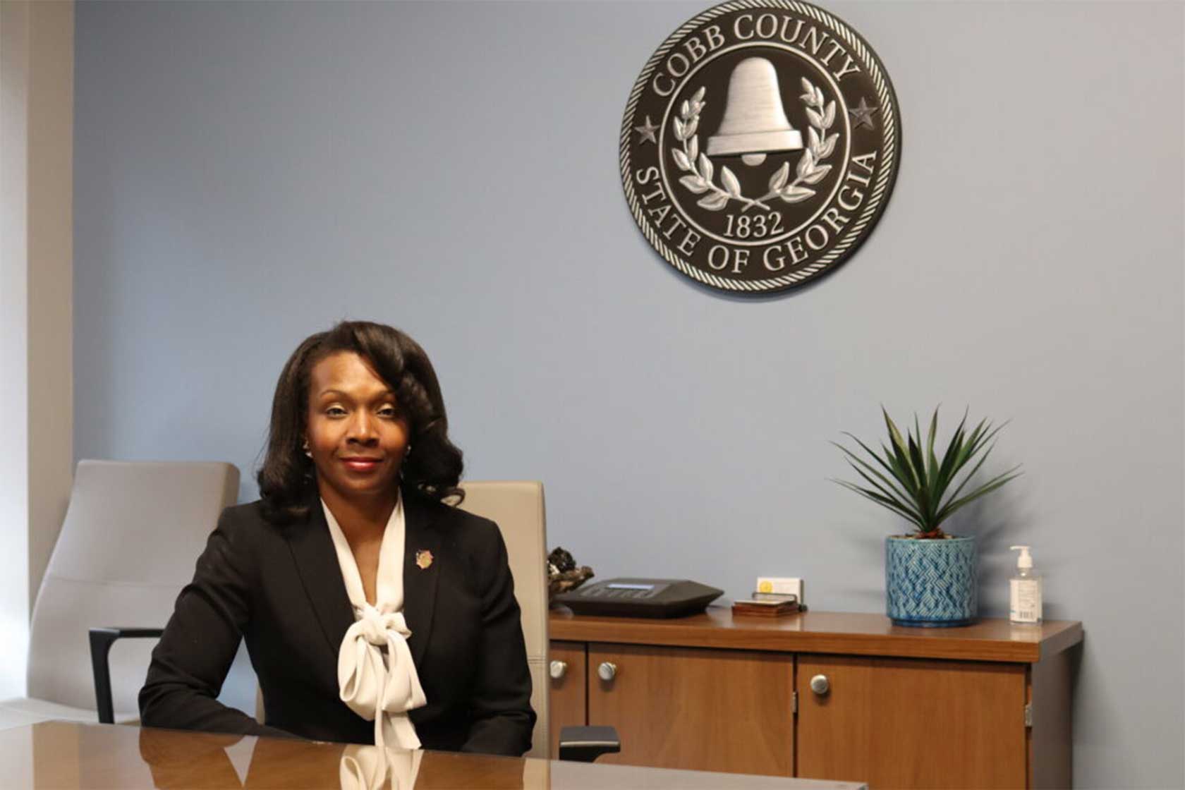A woman in a black blazer and white blouse sits at a desk in a professional office, with a blue wall and a Cobb County emblem behind her.
