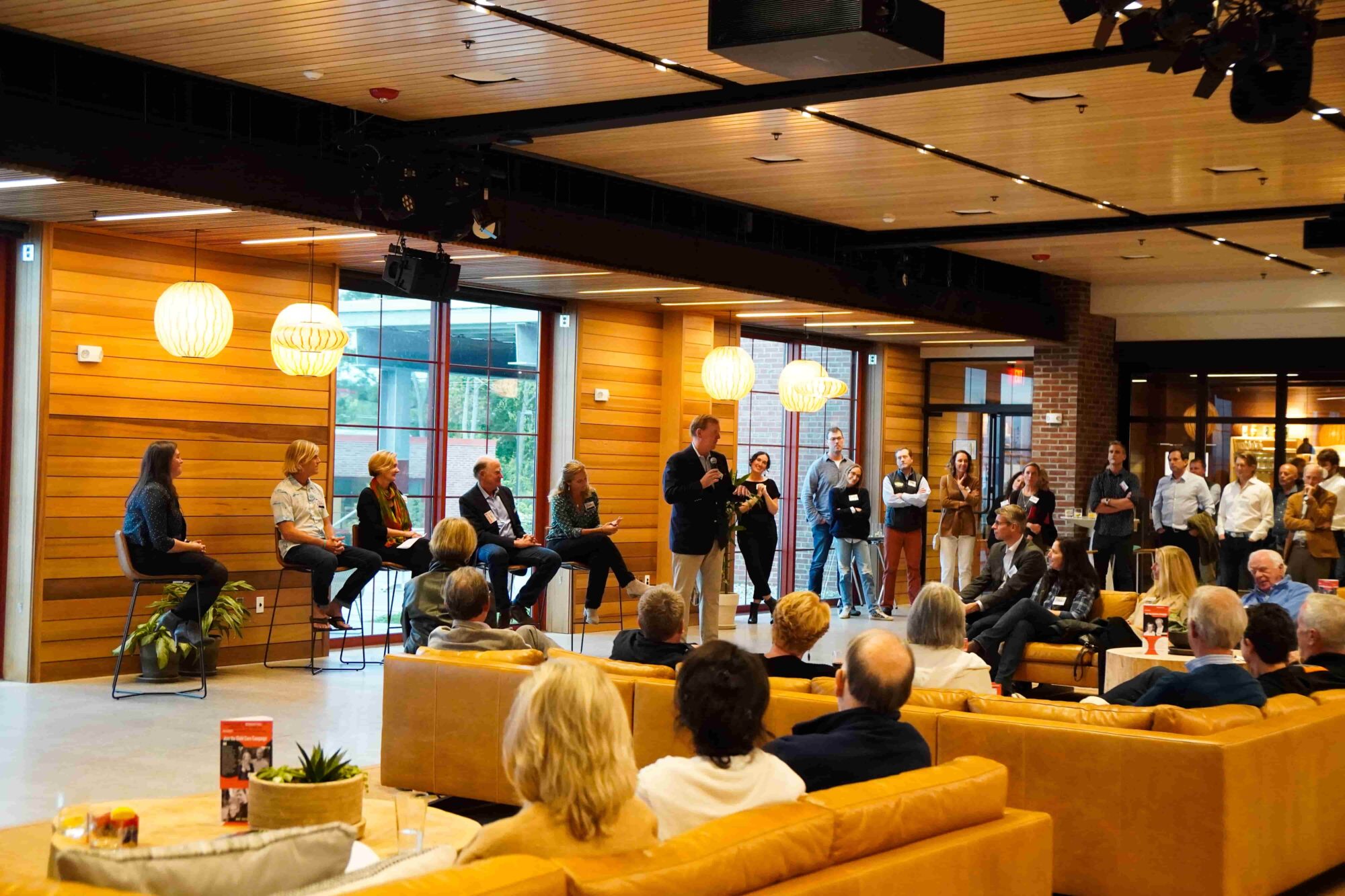 A panel discussion in a modern, wood-paneled room with attendees seated on couches and standing.