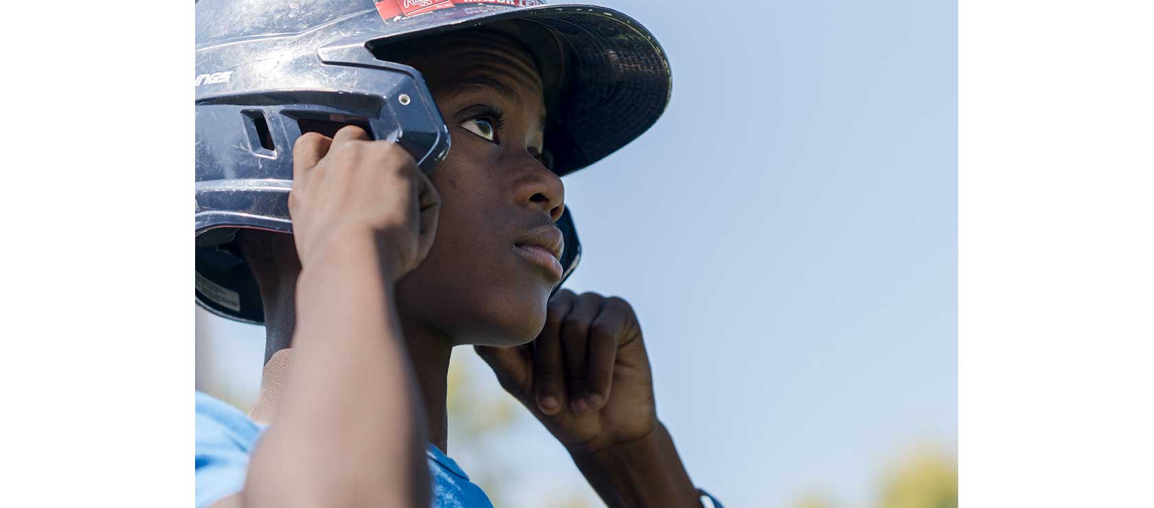 A young boy adjusts a black sports helmet while standing outdoors under a blue sky.