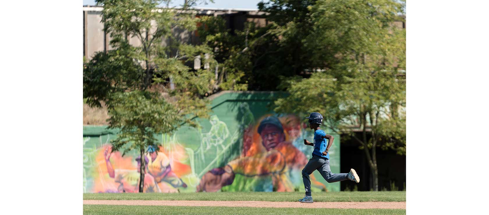 A young boy in a blue baseball jersey and helmet runs on a baseball field, with a colorful mural depicting baseball scenes in the background.