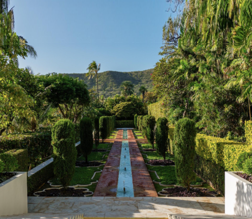 Vibrant green trees and shrubs line a long and narrow fountain pool. In the background is a rolling green hill with blue sky above.