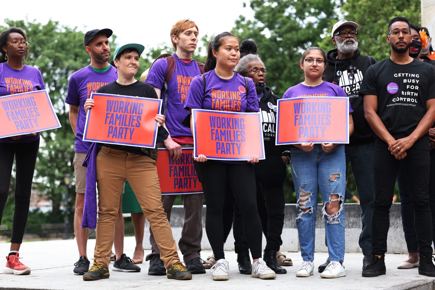 A diverse group of people, some wearing purple and orange shirts, hold matching signs for the Working Families Party.