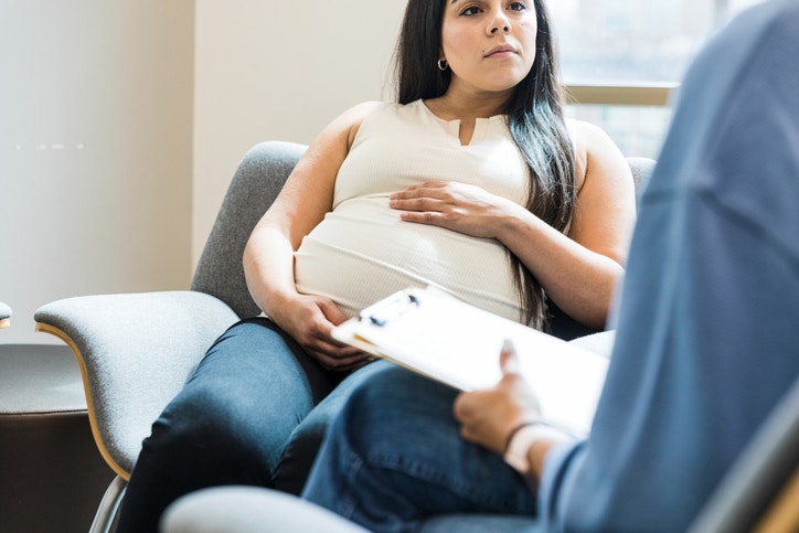 A pregnant woman sits on a chair listening to a person holding a clipboard.