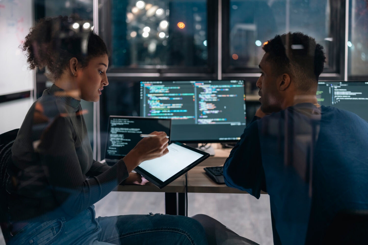 A casually dressed woman and man sit in an office with large windows at night. On the desk in front of them are monitors with code displayed. The woman holds a tablet and stylus as they hold discussion.