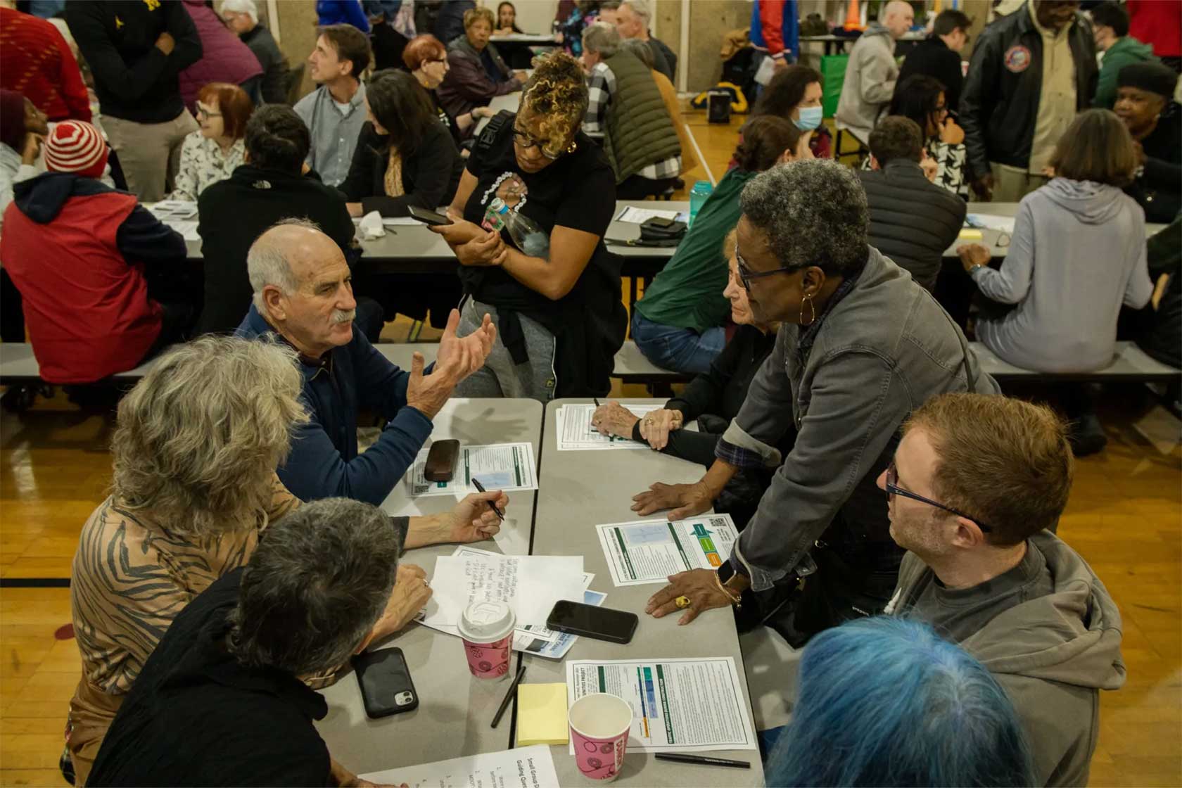 A community gathering in a gym with individuals engaged in conversations around tables, discussing various materials.