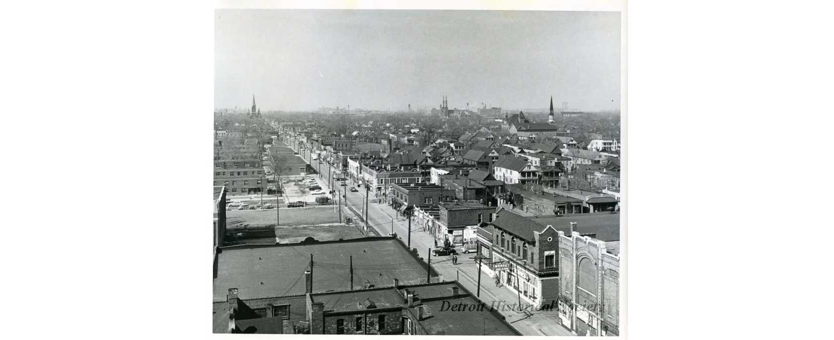 A black-and-white aerial view of a city, featuring residential houses, streets, and church spires in the background, with an overcast sky.