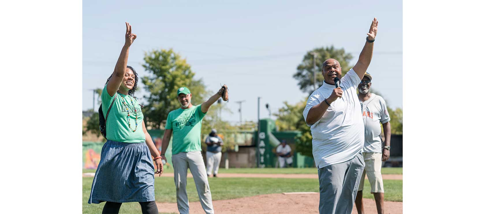 A group of three individuals stand on a baseball field, engaging in conversation during a sunny day. One woman gestures with her hand, while another man speaks into a microphone.