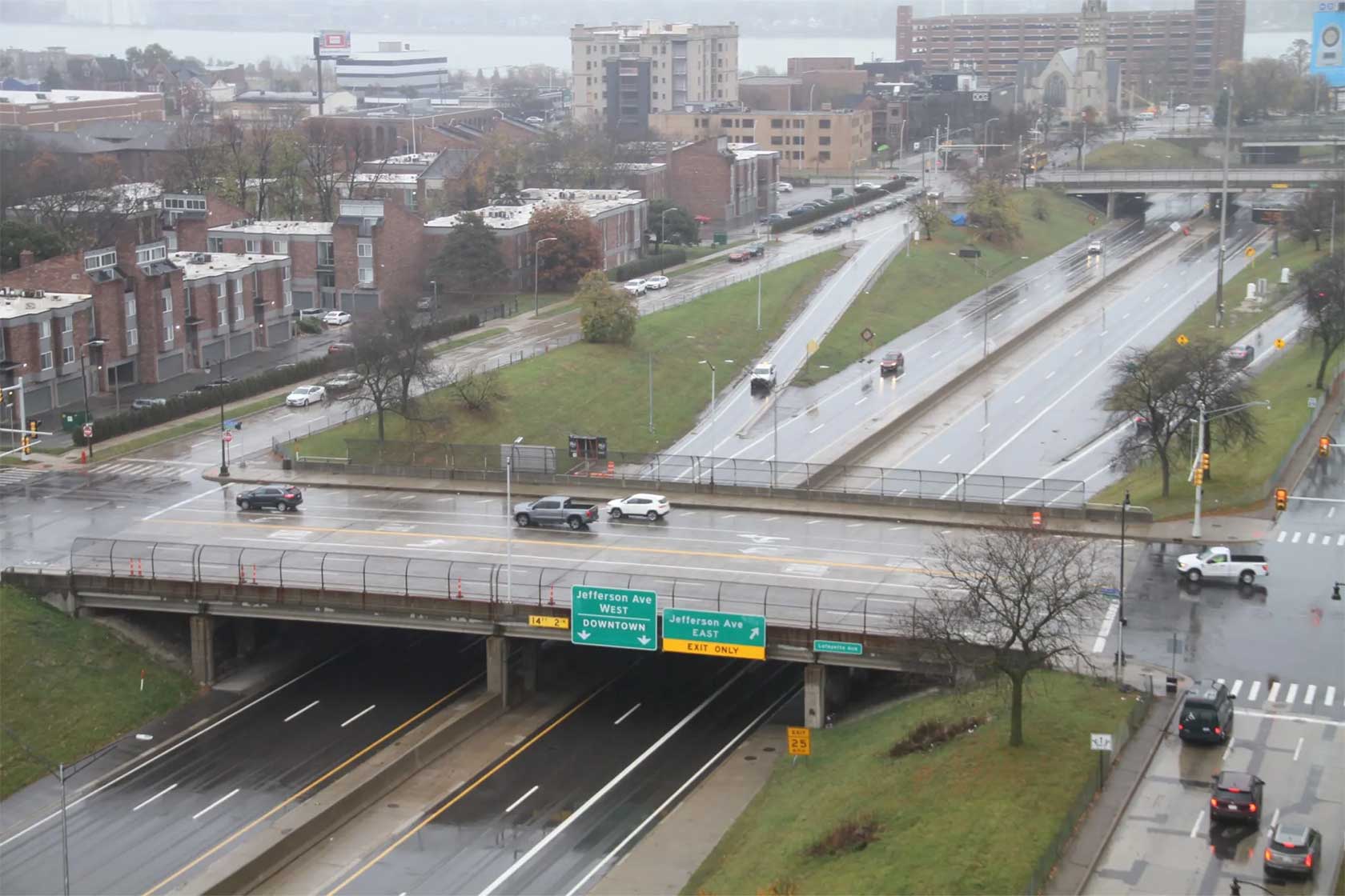 A rainy urban highway interchange showing an overpass with traffic signs directing to Jefferson Ave and Downtown, surrounded by residential buildings and a church in the background.