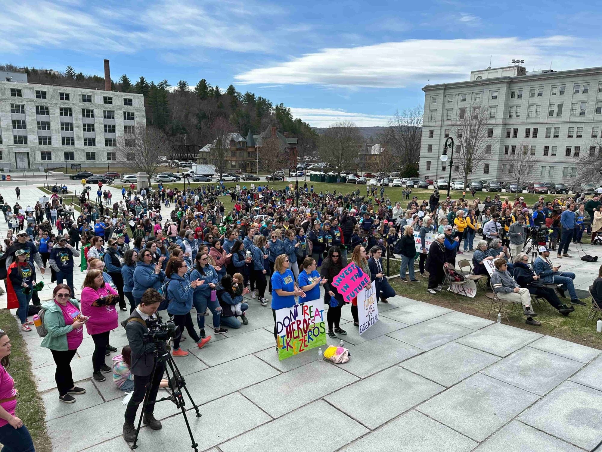 Crowd gathered on a stone platform outside government buildings, some holding signs.