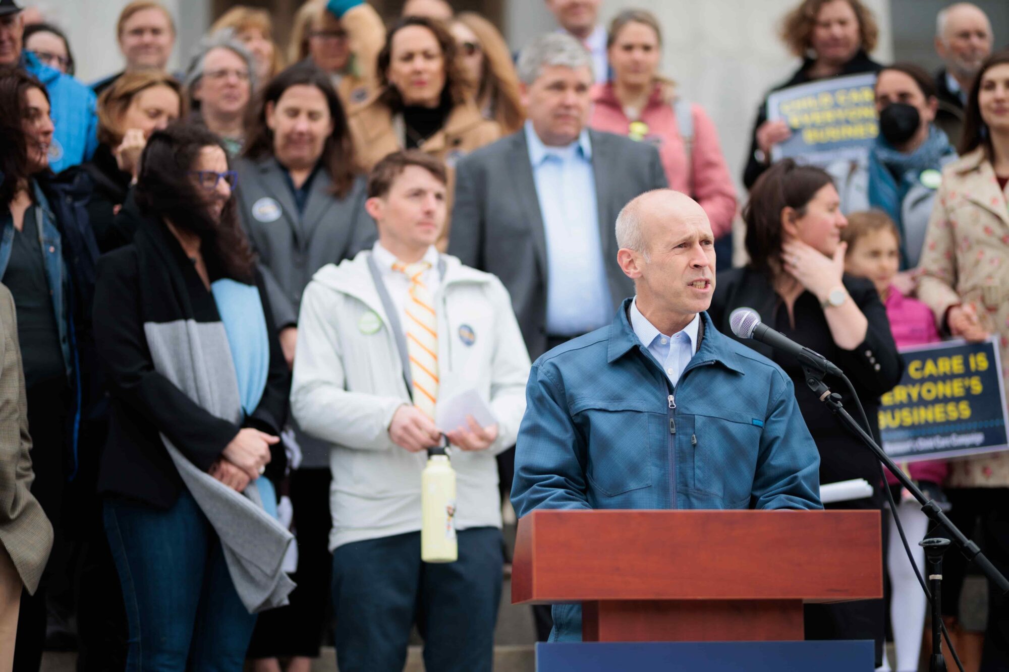 Person speaking at a podium in front of a group, some holding signs about child care.