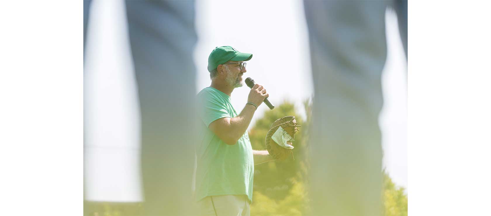 Man in a green shirt and cap speaking into a microphone outdoors while holding a baseball glove.