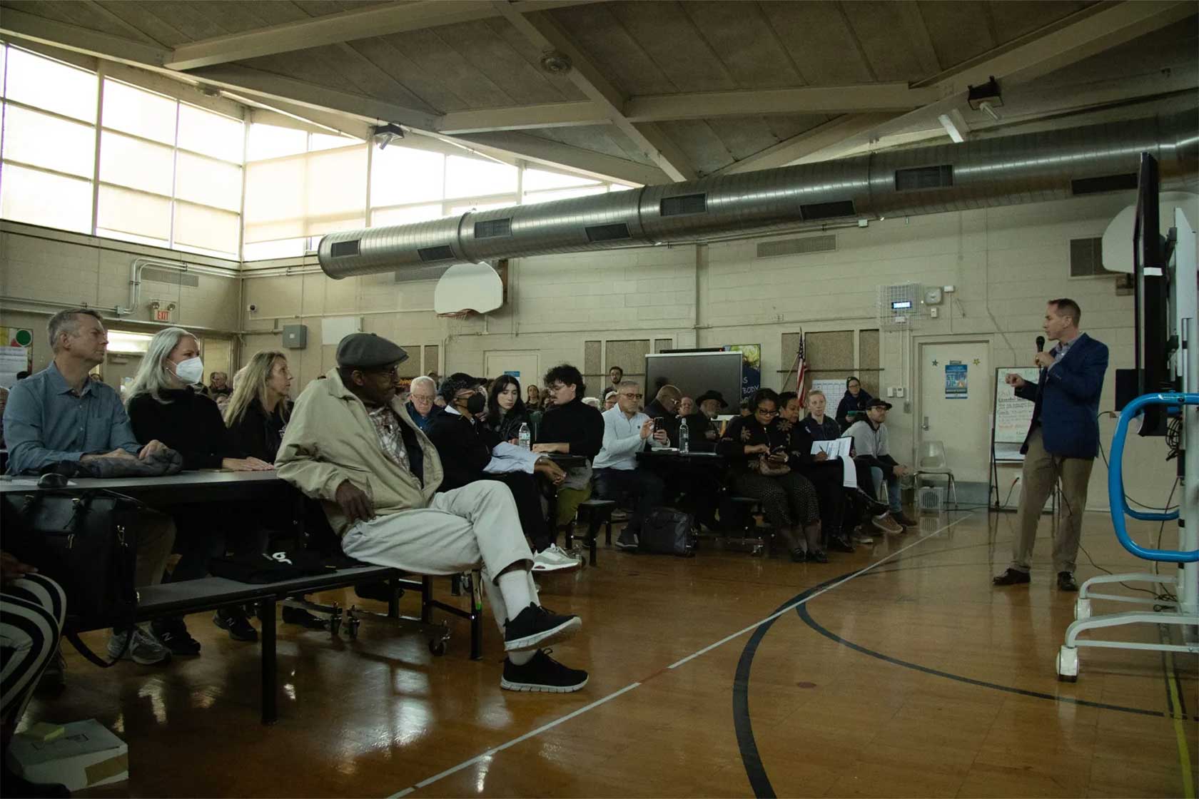A speaker in a blue jacket addresses a diverse audience seated on benches in a community center. Natural light streams through large windows, illuminating the gathering.