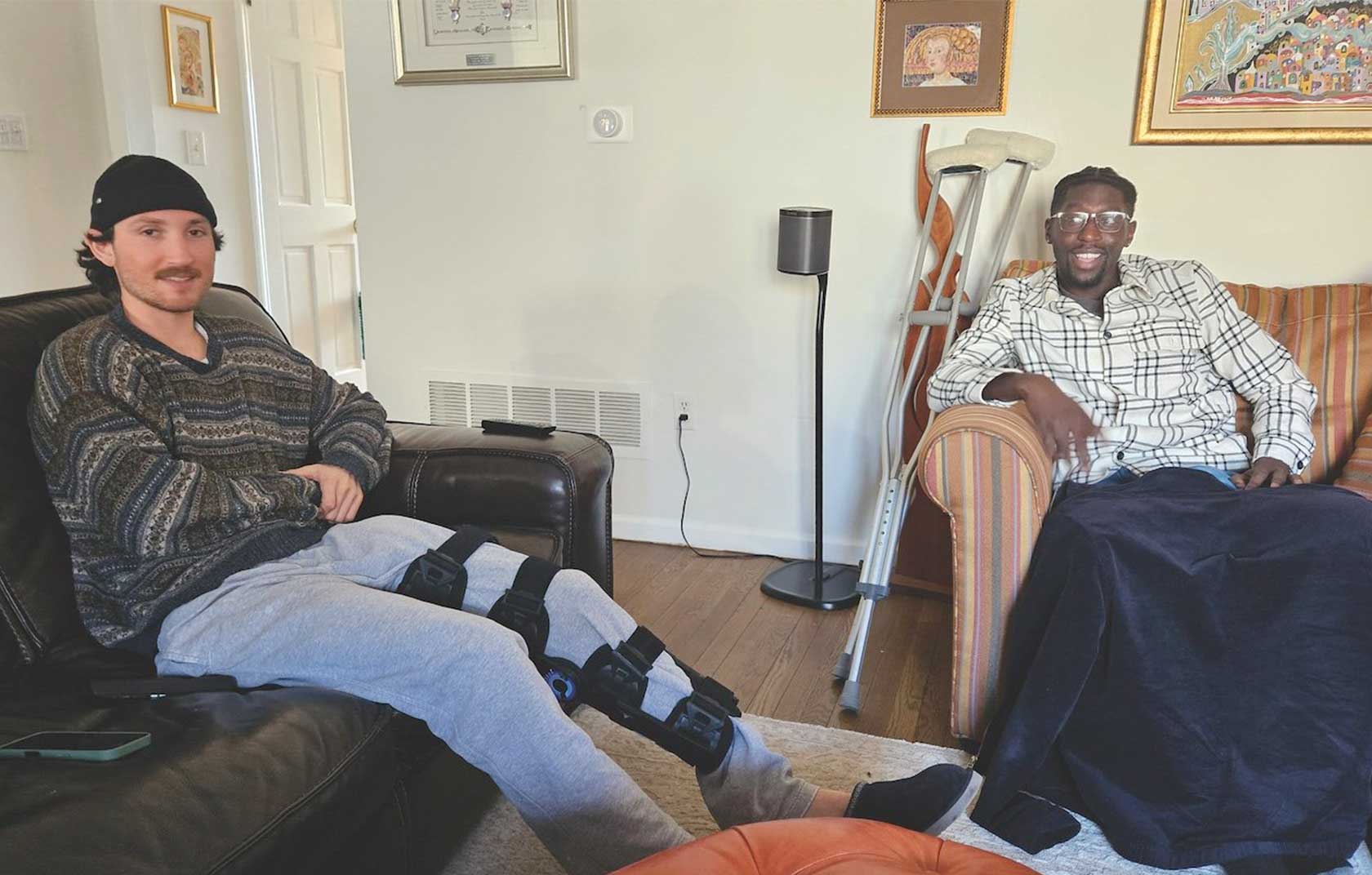Two young men sitting in a living room, one on a leather couch with a knee brace and the other on a striped chair with a navy blanket, both smiling.