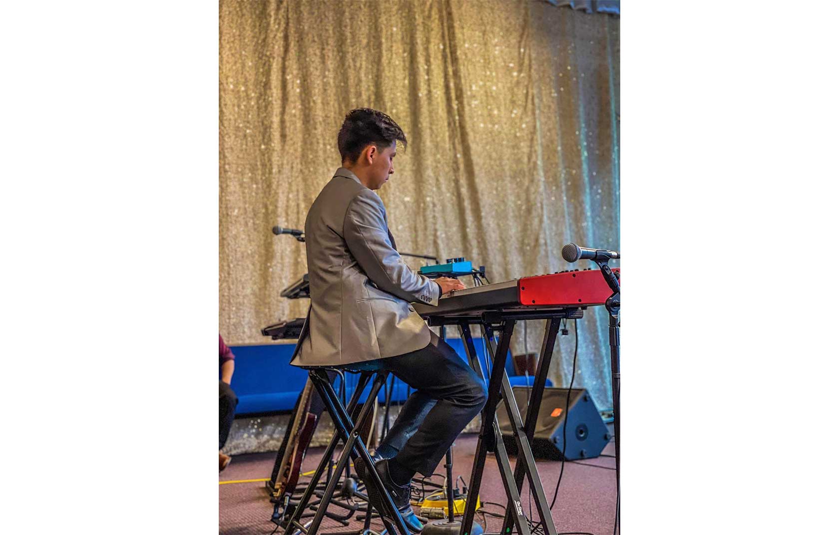 A young man in a gray suit plays an electronic keyboard on stage against a sparkling gold backdrop.