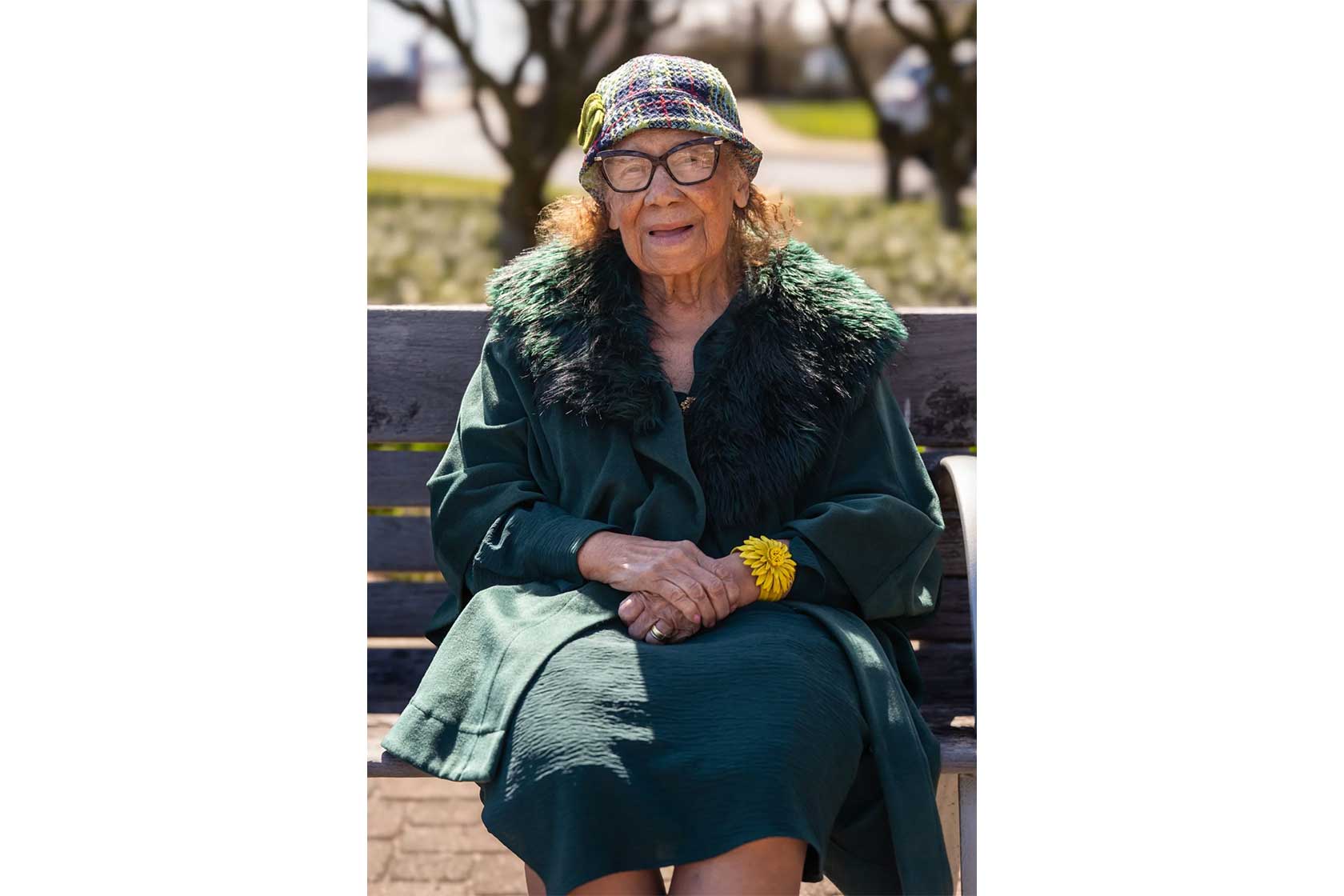 An elderly woman in a green dress with a fur collar sits on a bench, wearing glasses and a yellow flower bracelet.