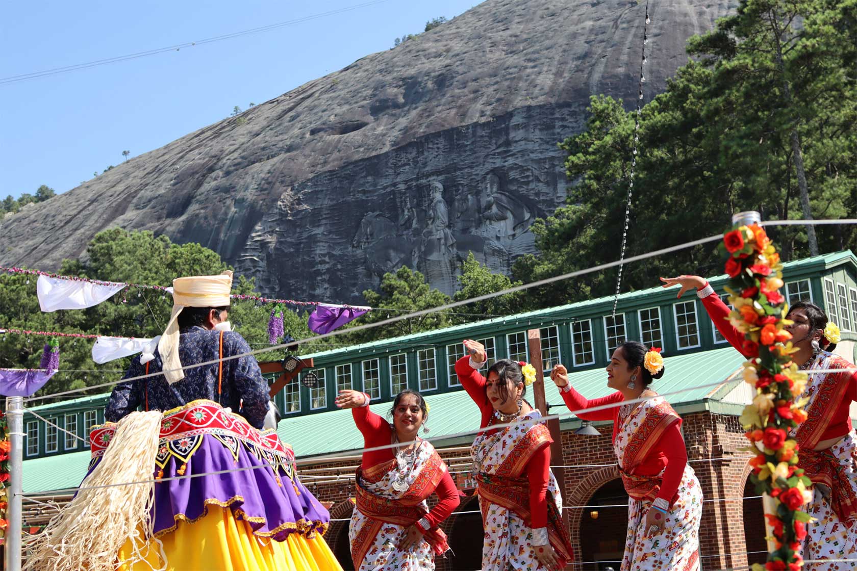 A performer in a traditional outfit stands in front of four dancers in white saris, dancing joyfully in a colorful outdoor cultural event with a large rock formation in the background.