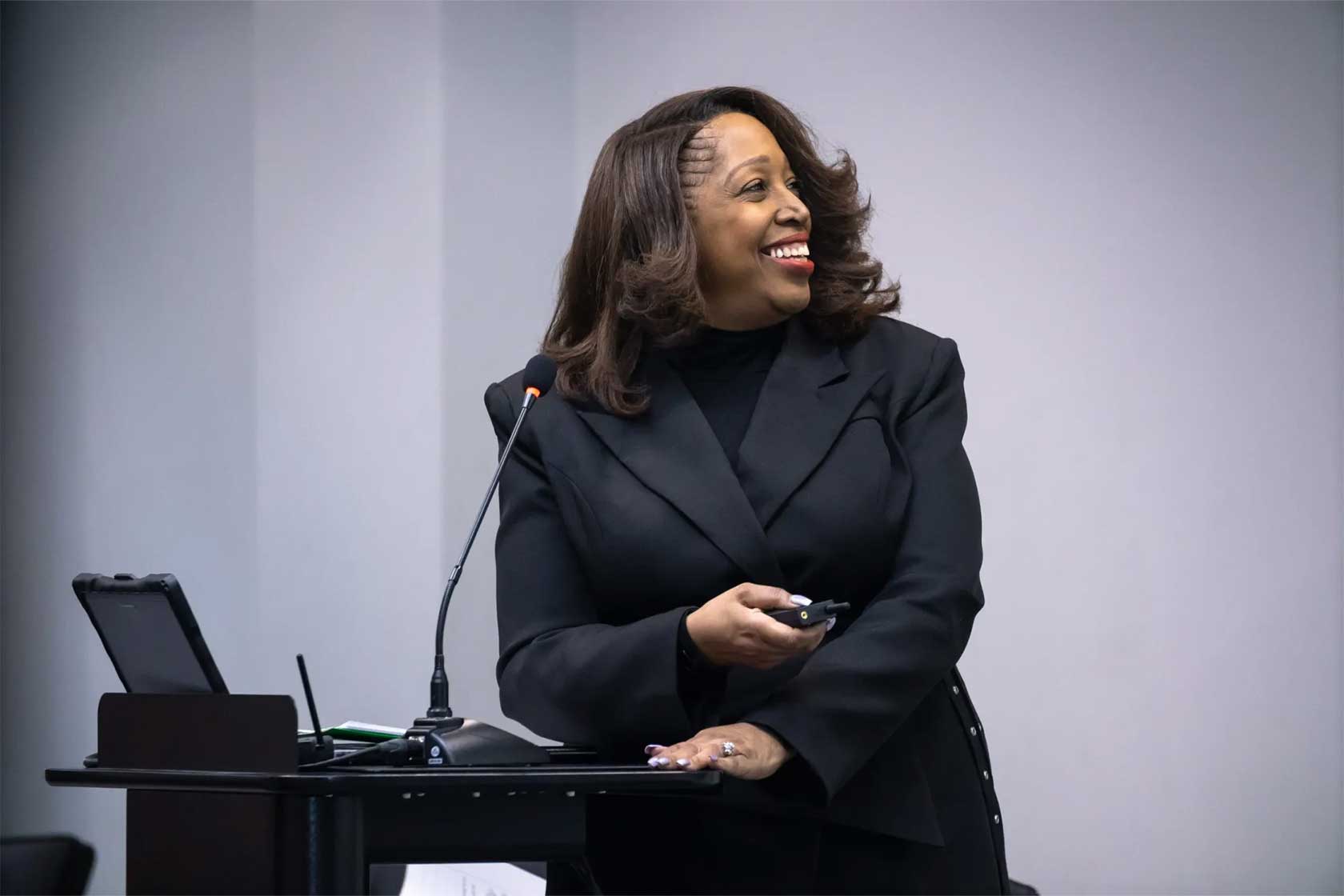 A smiling woman in a black suit stands at a podium, gesturing with one hand while holding a presentation remote in the other.