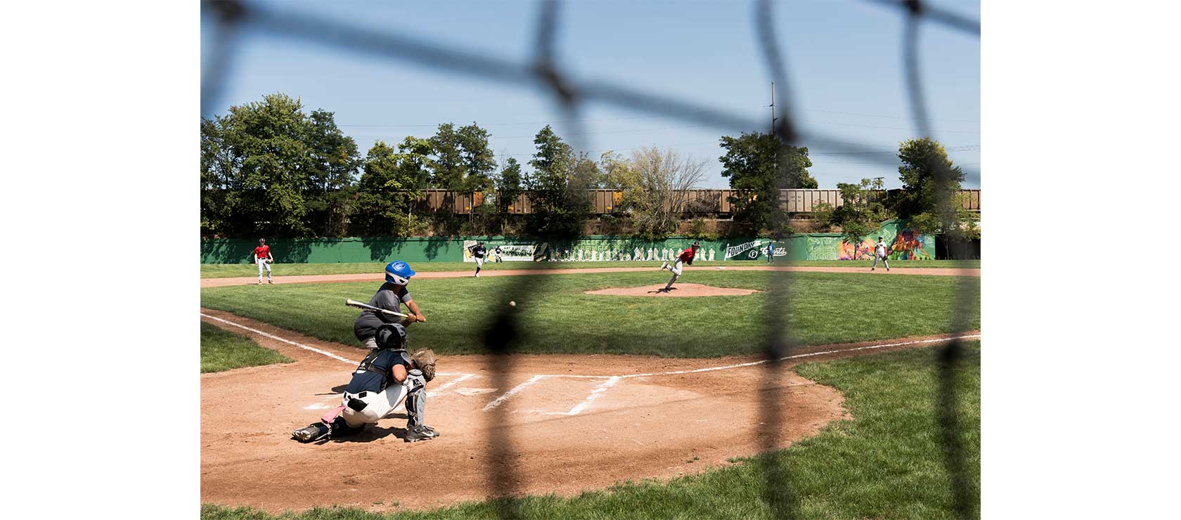 A youth baseball game with a catcher in black and white crouching behind home plate while a pitcher in red throws the ball to a batter, framed by a chain-link fence.