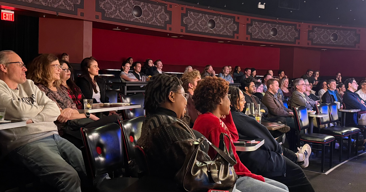 A diverse audience sits attentively in a vibrant, vintage comedy venue, engaged and smiling, with drinks on tables in front.