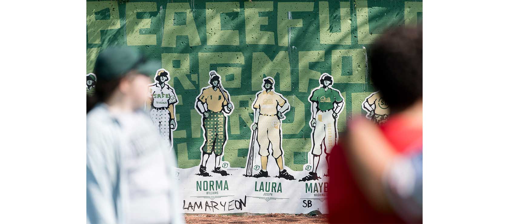 Mural of four women in vintage baseball uniforms on a green wall, with the word “PEACEFUL” in the background. Names under the figures include 