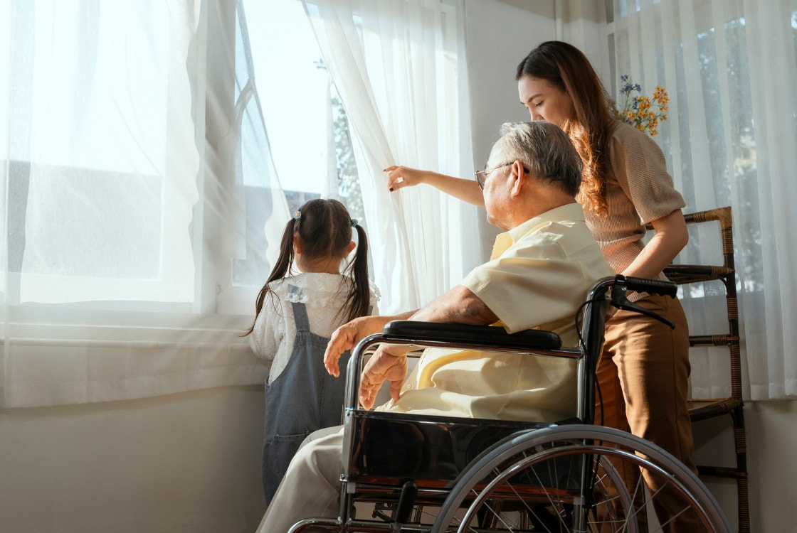 An adult woman gestures toward a window where she stands next to an older man in a wheelchair and a young girl with pigtails.