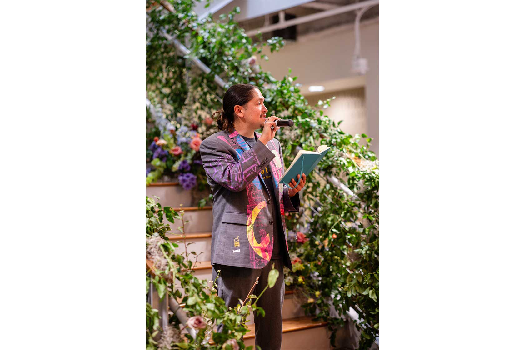 A man in a colorful blazer stands on a staircase, holding a microphone and an open book, surrounded by lush greenery and flowers.
