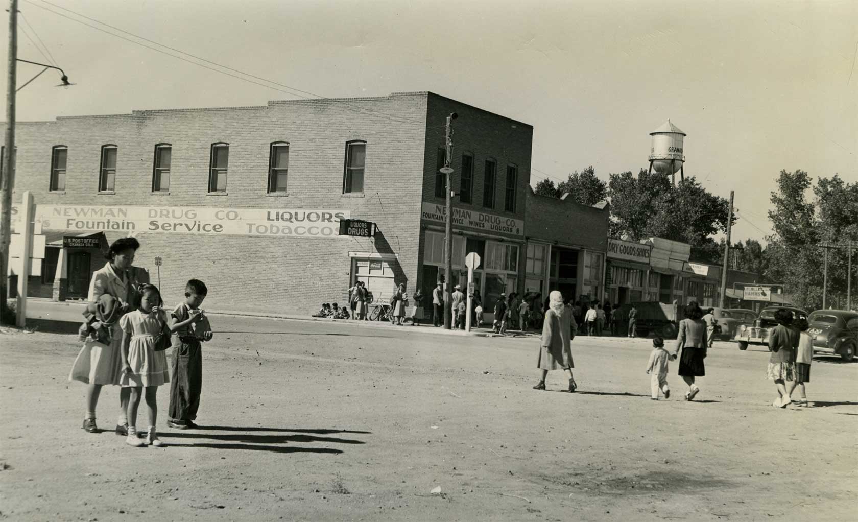 A historic street scene with children in the foreground and several businesses in the background, including a drug store and a dry goods shop. A water tower is visible in the distance.