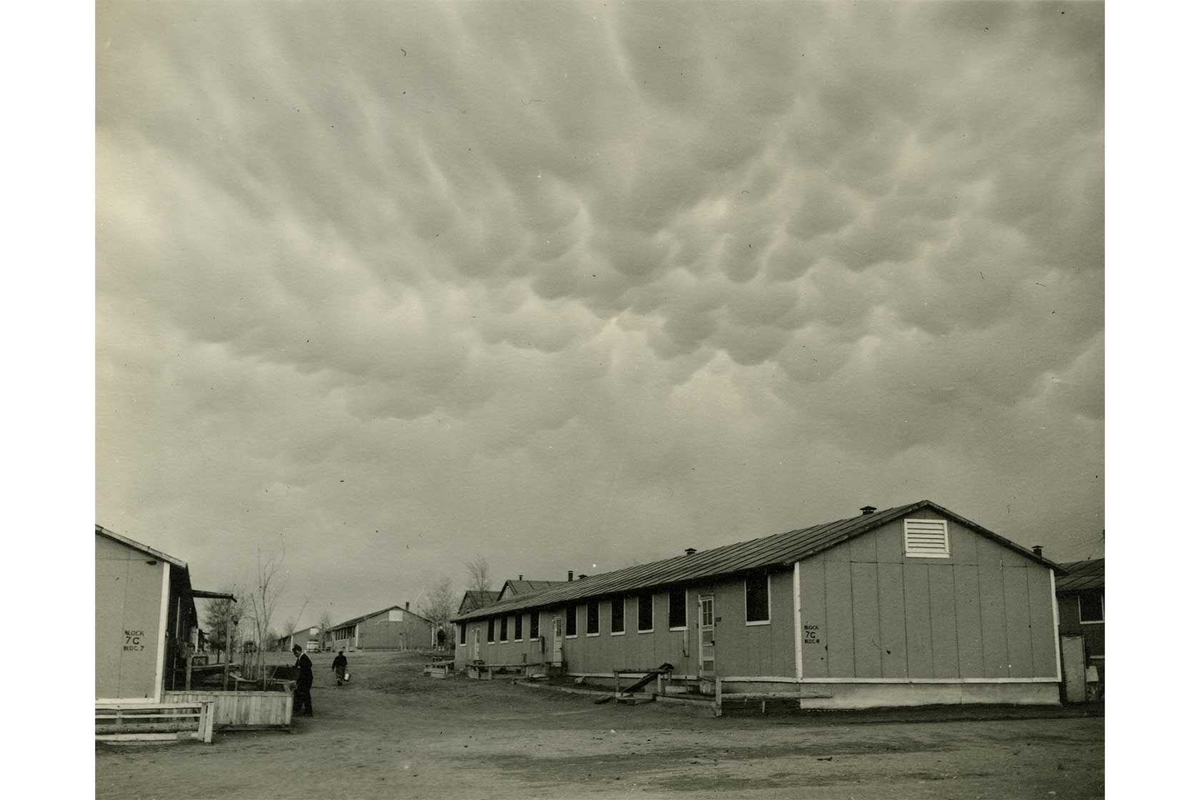 A row of simple buildings under a textured, cloudy sky, with two people walking along a dirt path.