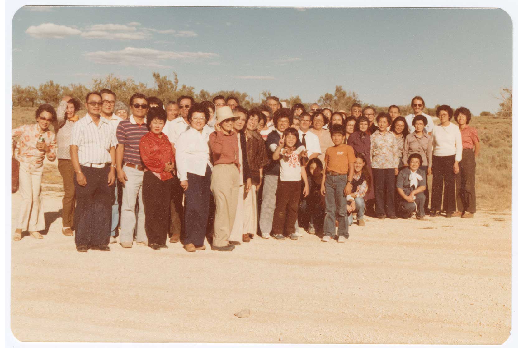 A large group of about 40 people stands together outdoors in a rural setting, smiling and posing for the camera, typical of the late 1970s style.