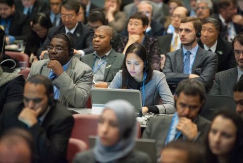 Attendees at a conference, including a woman using a laptop, surrounded by others wearing headphones.