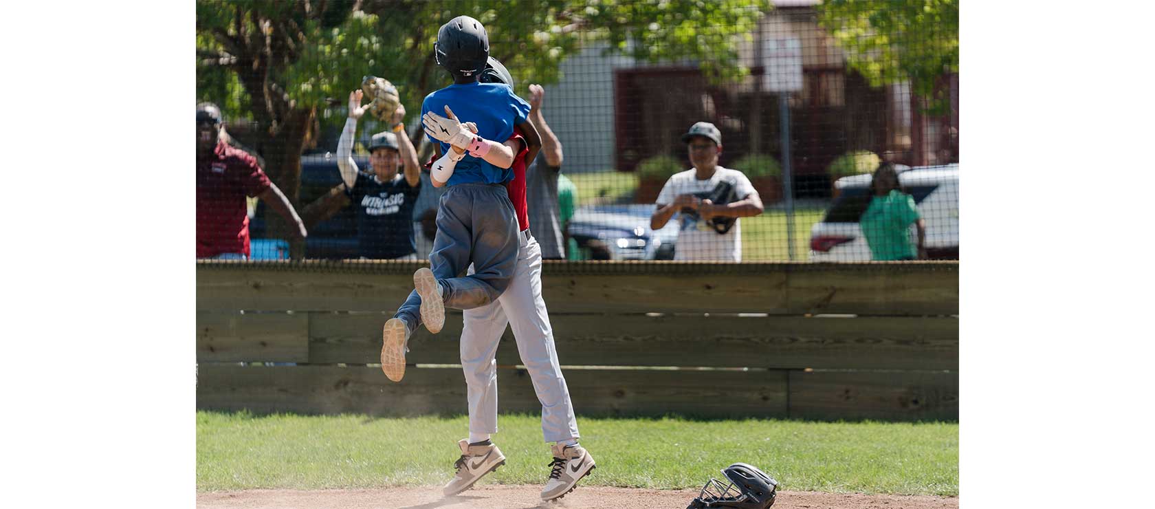 Two youth baseball players celebrate a victory by jumping into each other's arms, surrounded by cheering spectators at a baseball field.