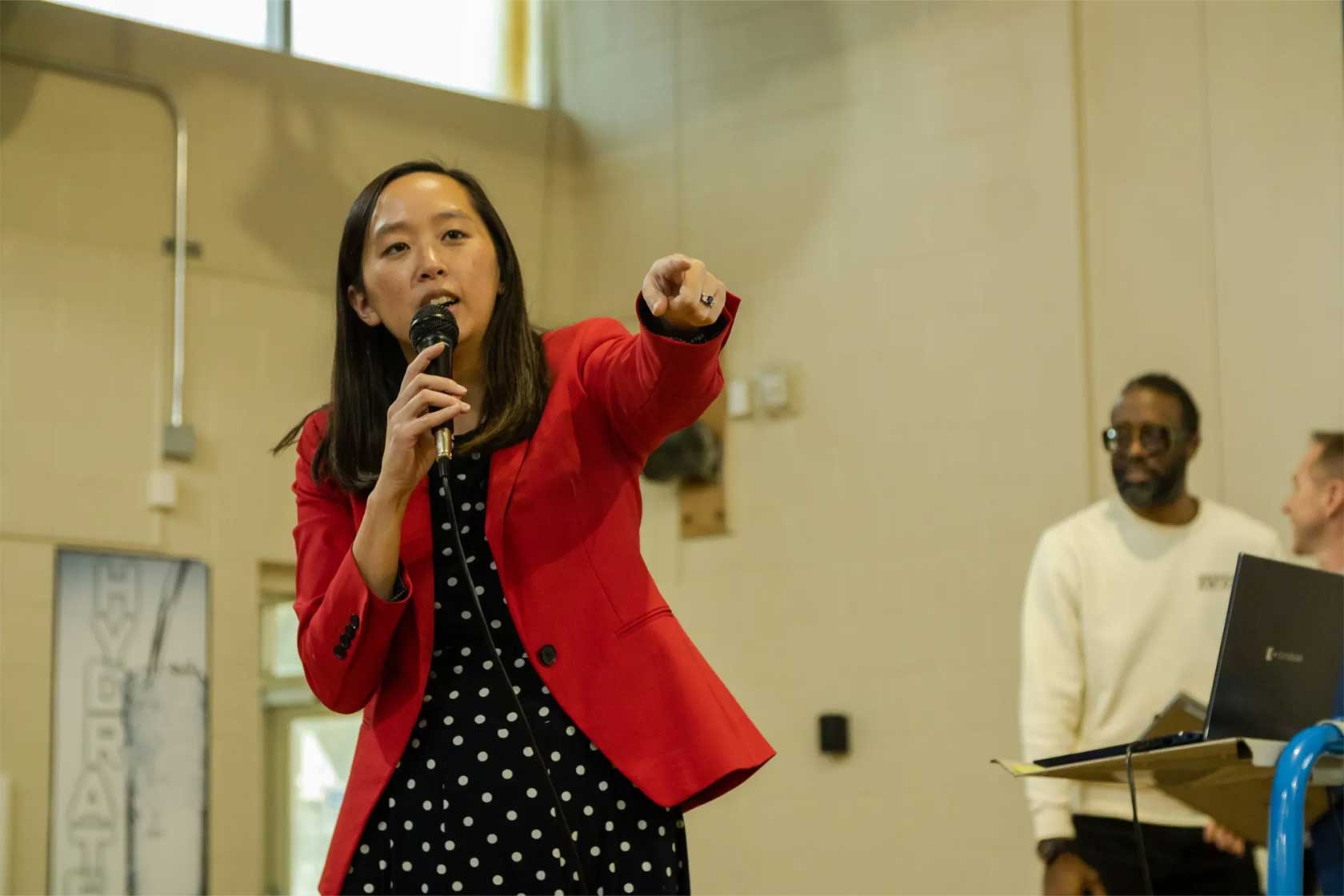 A woman in a red blazer and polka dot dress is speaking into a microphone, pointing with her left hand. A man in a white sweatshirt stands in the background, observing.