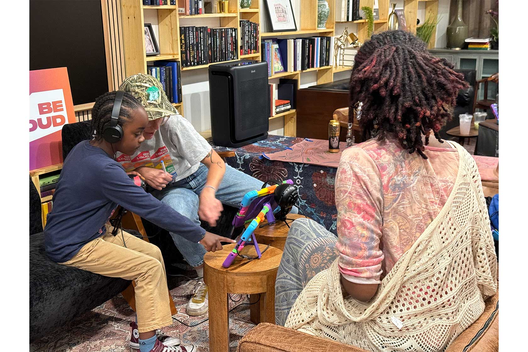 A girl in headphones interacts with a colorful device on a table, while a young adult in a camouflage hat assists her. A woman with curly hair observes from the side in a cozy living room.