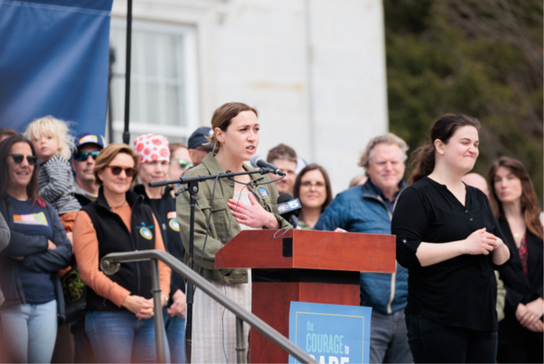 A woman speaks at a podium with microphones at an outdoor event, surrounded by a diverse group, including a sign language interpreter and other attendees.