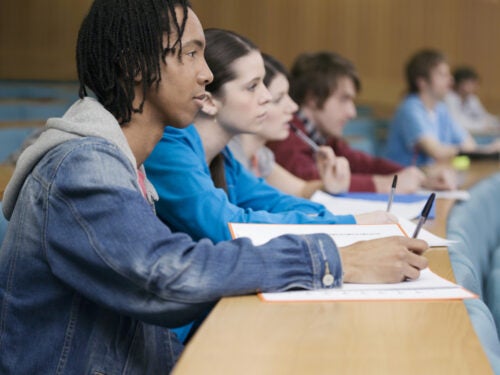 A row of college students sitting at desks in a lecture hall, taking notes and listening.
