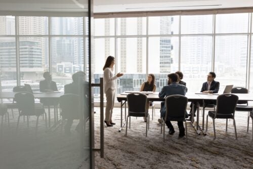 A group of 4 diverse business people sit at a table, listening to a woman standing at the front of the room.