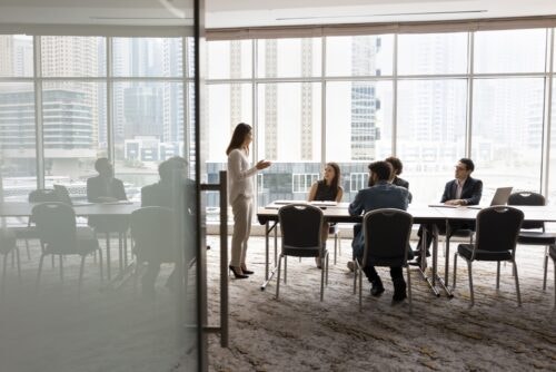A group of 4 diverse business people sit at a table, listening to a woman standing at the front of the room.