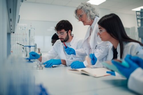 A group of two diverse students sit at a lab table with a professor guiding them.